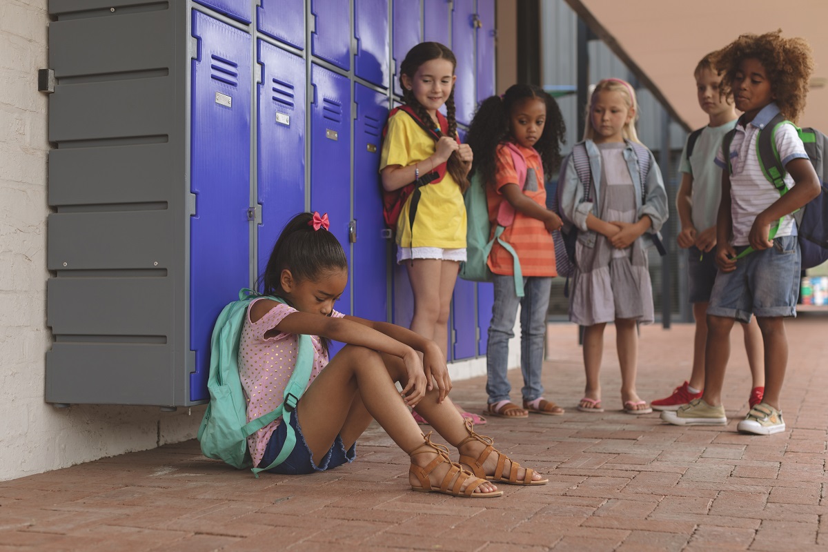 Side view of a schoolgirl sitting alone in school corridor while others school kids looking at her in background