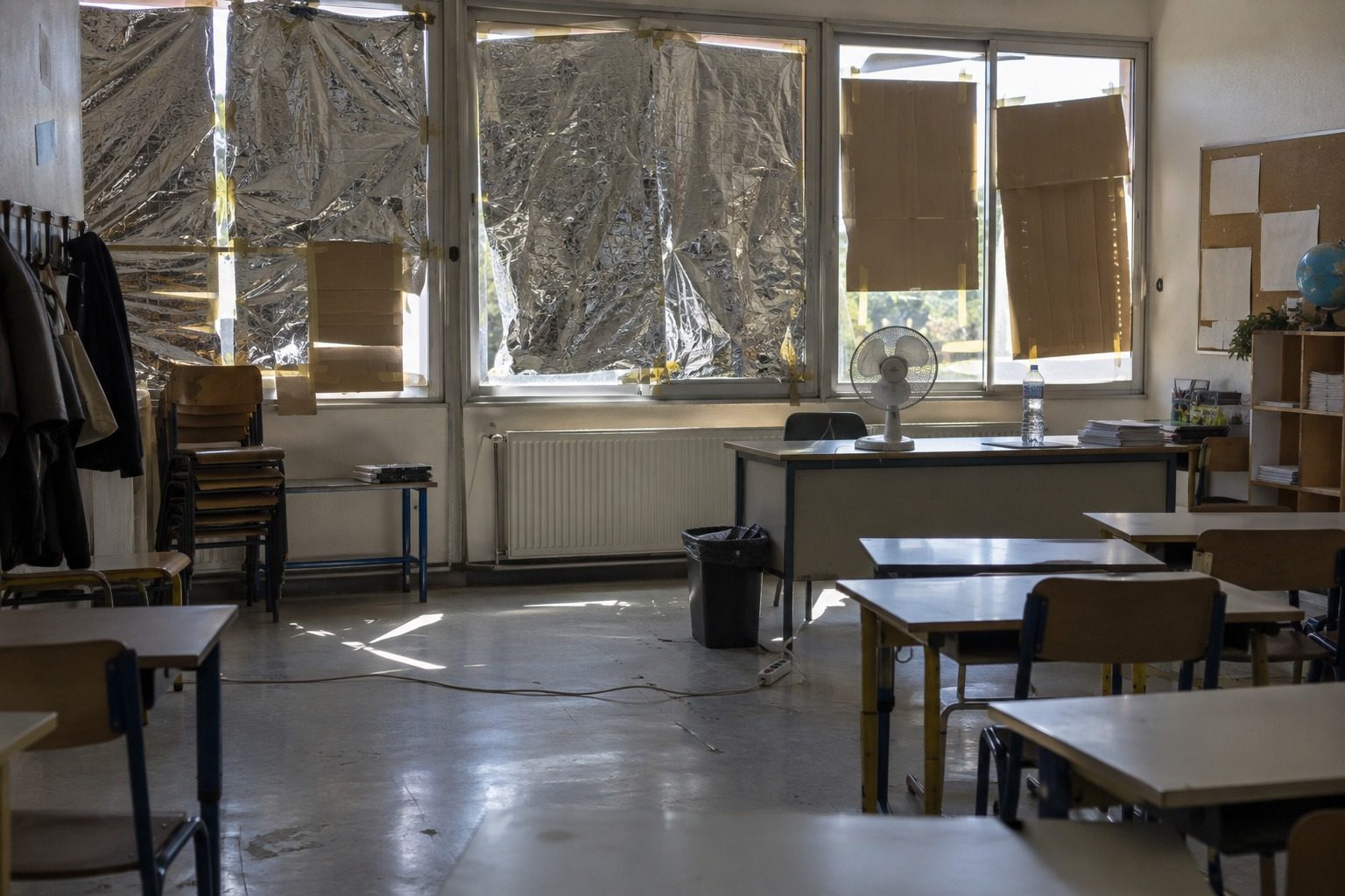 Salle de classe vide en pleine chaleur, fenêtres tapissées de couvertures de survie et petit ventilateur sur le bureau